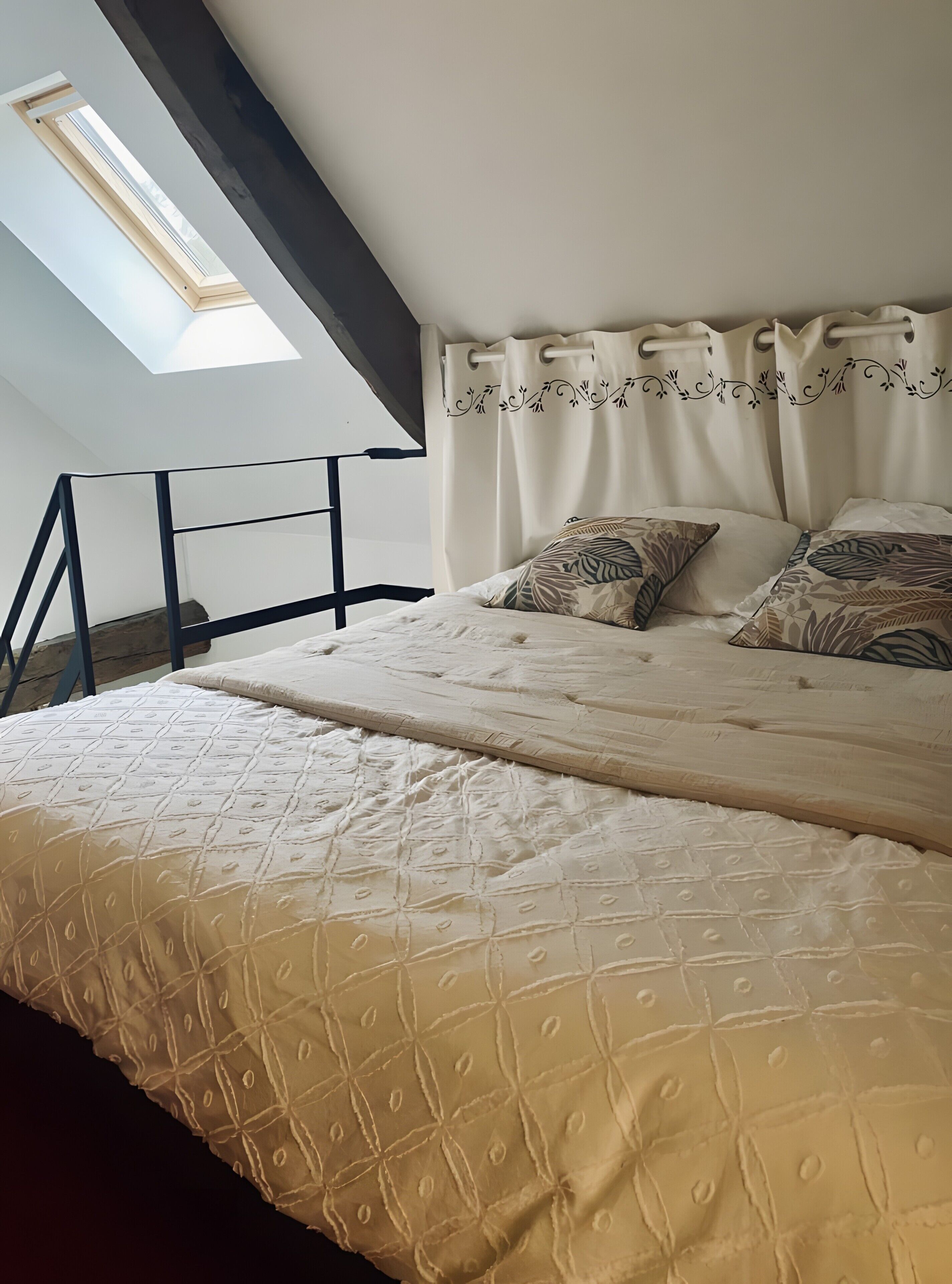 A bedroom with a skylight, a bed with patterned pillows, and a headboard with curtains.