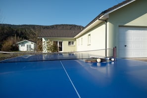 Een blauwe tafeltennistafel met net, een witte garagepoort, en een huis met een lichtgekleurde gevel en een donker dak.