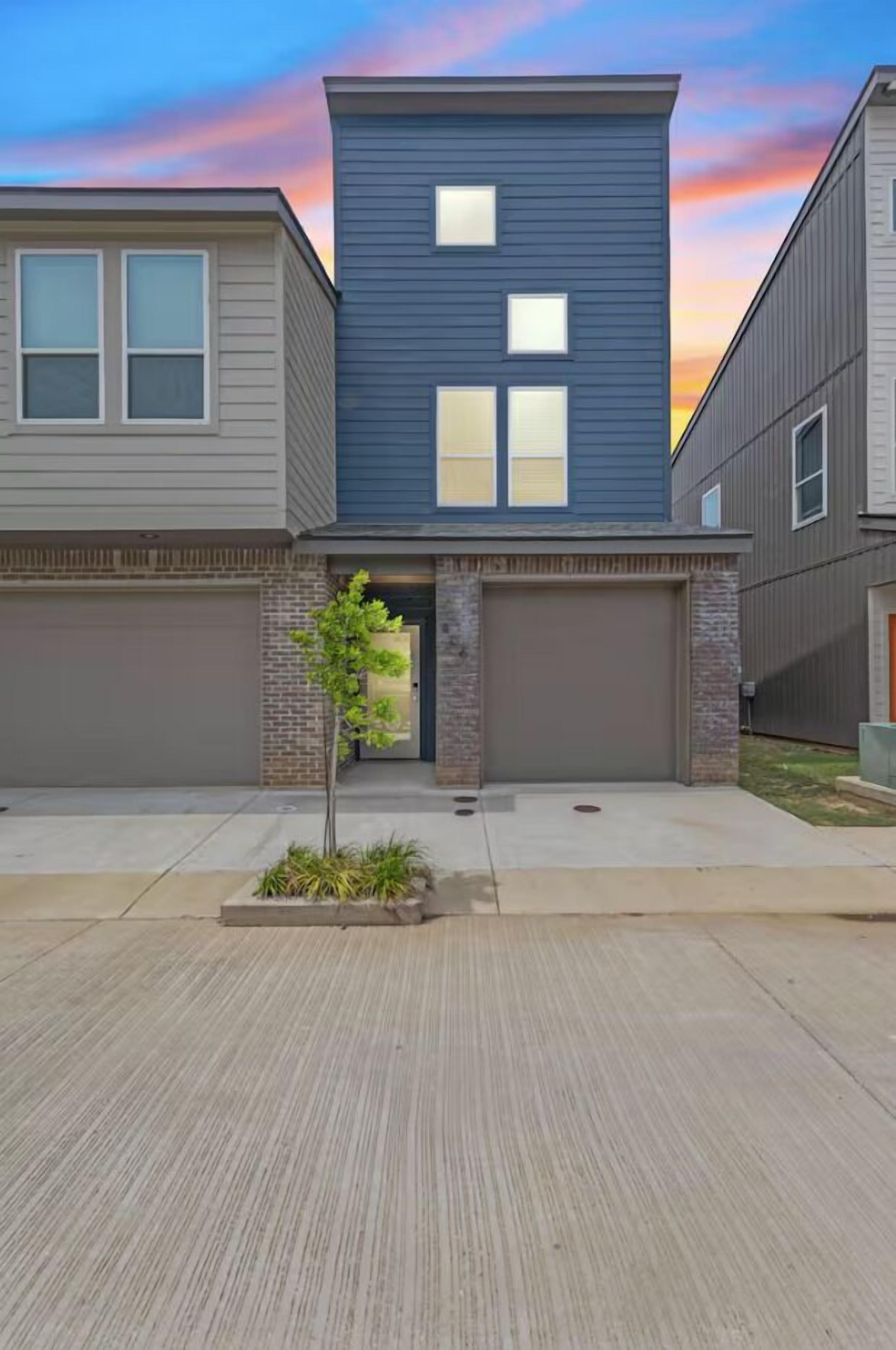 A modern two-story residential building with a garage, a small tree in the front yard, and a clear sky.