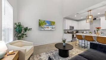 A modern living room with a sofa, a coffee table, and a TV. There is a kitchen in the background with white cabinets and a bar area.