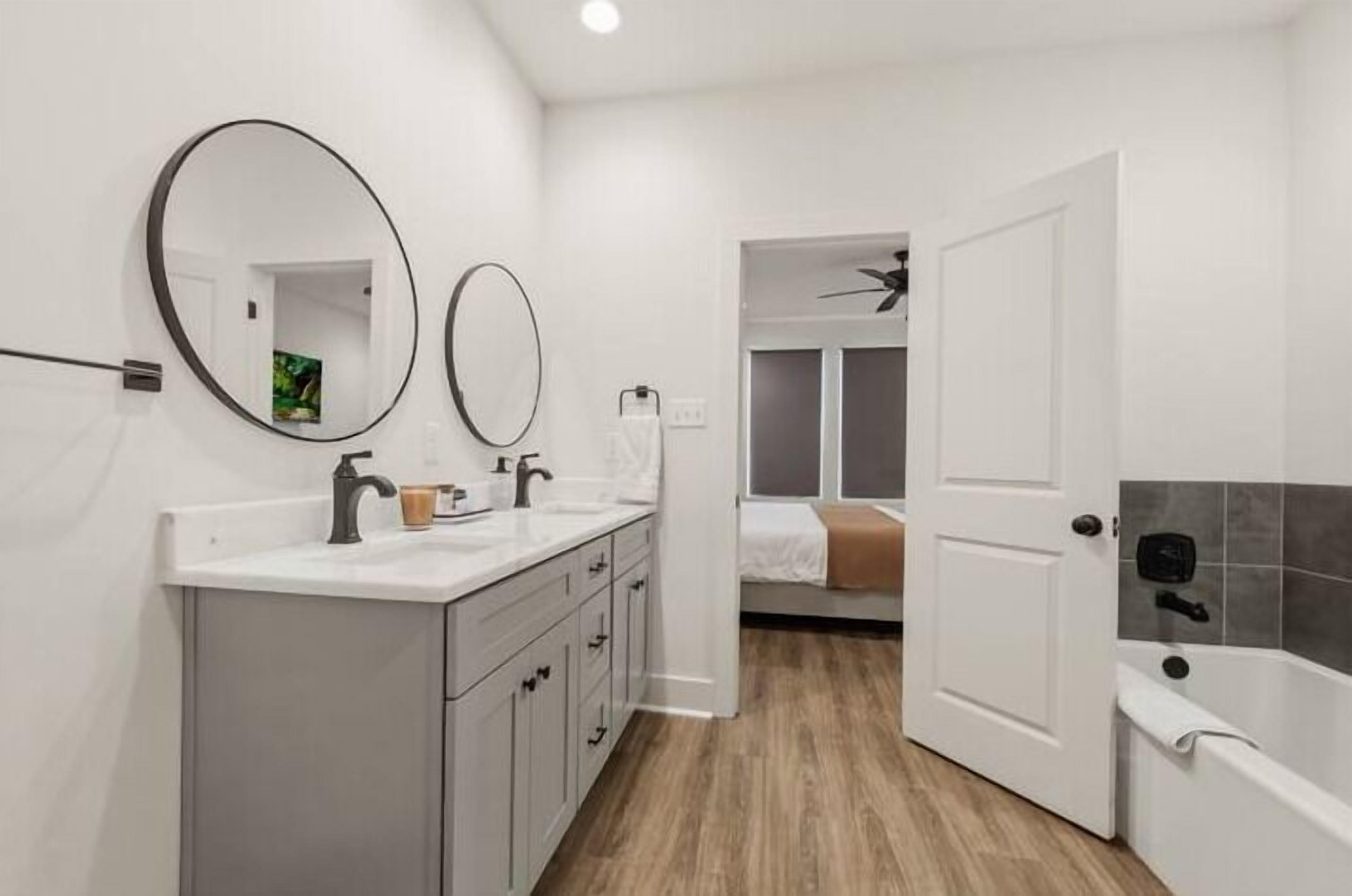 Un baño con un lavamanos blanco, dos espejos, una bañera y una puerta que da a un dormitorio.