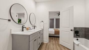 Un baño con un lavamanos blanco, dos espejos, una bañera y una puerta que da a un dormitorio.