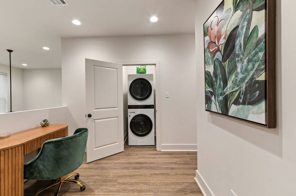 Un cuarto de lavandería con dos lavadoras y secadoras apiladas, un escritorio de madera con una silla verde, y un cuadro grande con motivos florales en la pared.