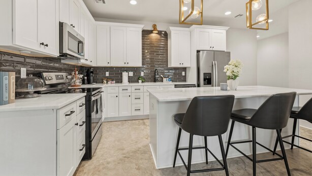 A modern kitchen with white cabinets, a central island with bar stools, a stainless steel refrigerator, and a brick backsplash. - Enclave 2 Bedroom Luxury Homes (Lafayette)