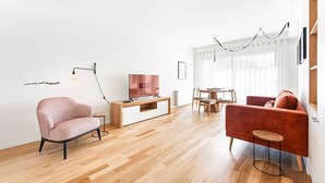 Un salon moderne avec un plancher en bois, un canapé rouge, un fauteuil rose, un téléviseur posé sur un meuble en bois, et un coin repas avec une table et des chaises.