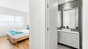 Une chambre d’hôtel moderne avec un lit en bois, un grand miroir et une salle de bain dont le mur derrière le lavabo est recouvert de carreaux.