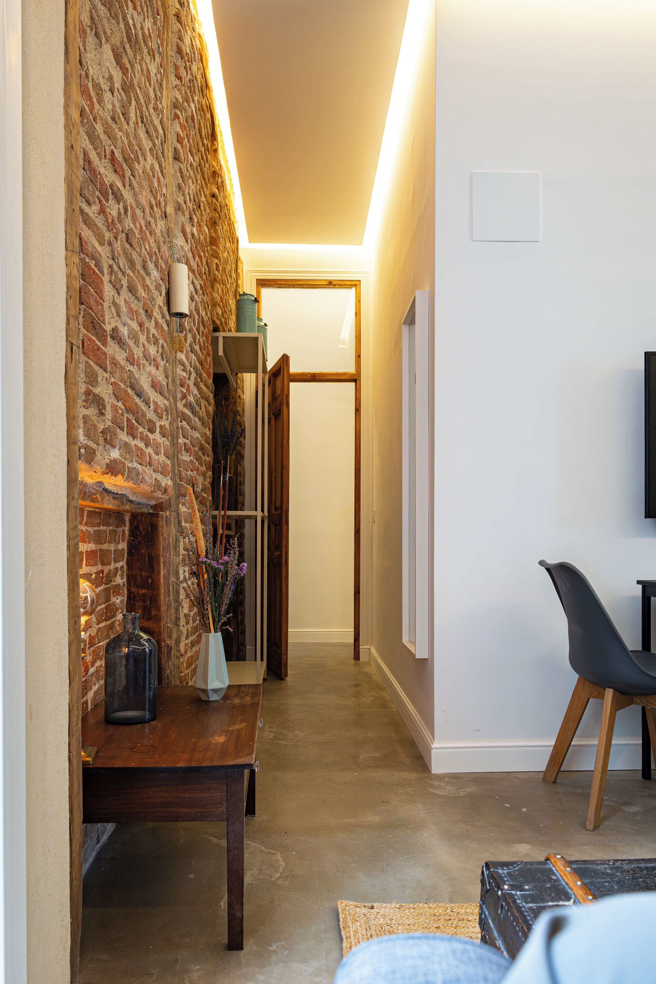 Un pasillo estrecho con una pared de ladrillo a un lado y una pared blanca al otro, que conduce a una puerta. Una mesa de madera con un florero y una lámpara está colocada contra la pared de ladrillo.