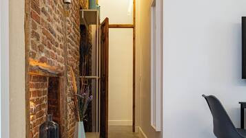 Un pasillo estrecho con una pared de ladrillo a un lado y una pared blanca al otro, que conduce a una puerta. Una mesa de madera con un florero y una lámpara está colocada contra la pared de ladrillo.
