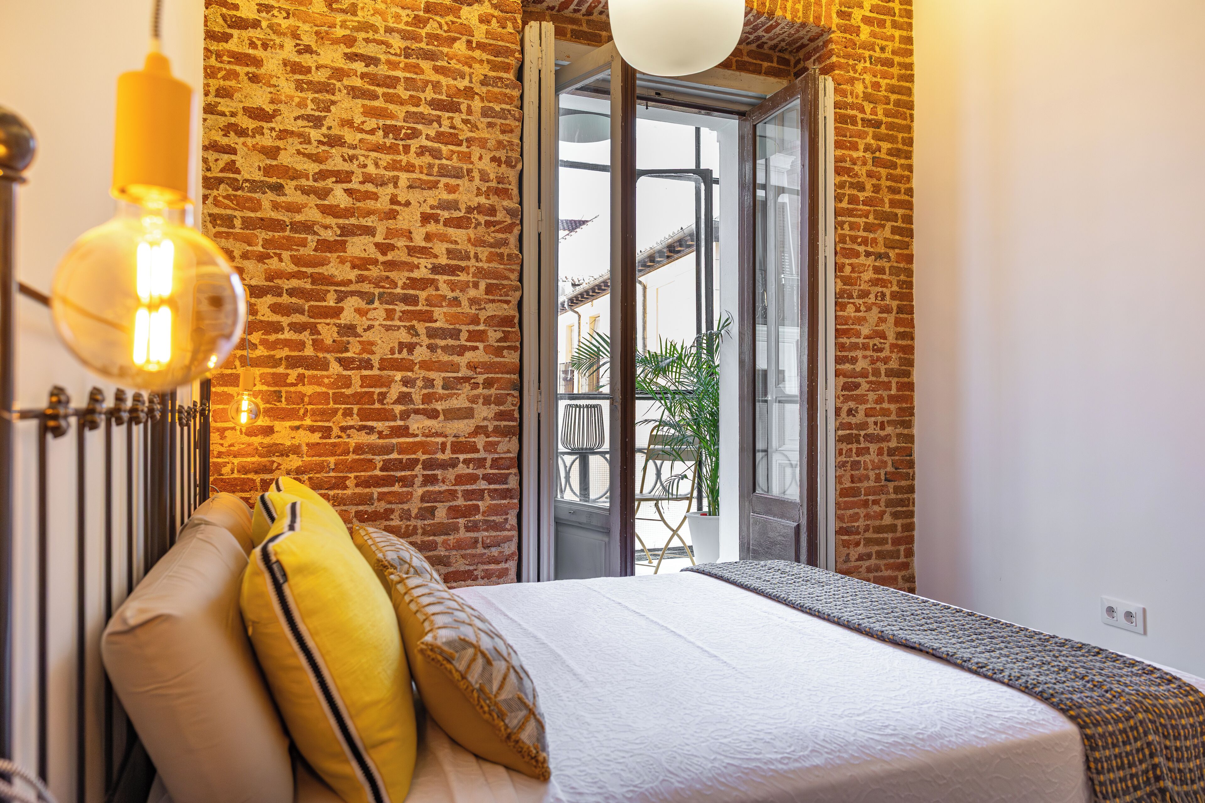 Un dormitorio con pared de ladrillo, cama con almohadas amarillas y beige, lámpara colgante y vistas a través de un ventanal.