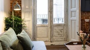 A room with a brick wall, a white door with a balcony, a grey sofa, a small table with a vase, and a floor lamp. - Uma Suites Fuencarral (Madrid)