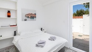 A bedroom with a bed, white walls, a picture of a coastal town, a white door, and a small shelf with a bottle. - Sunny Palace Studio 2 (Dubrovnik)