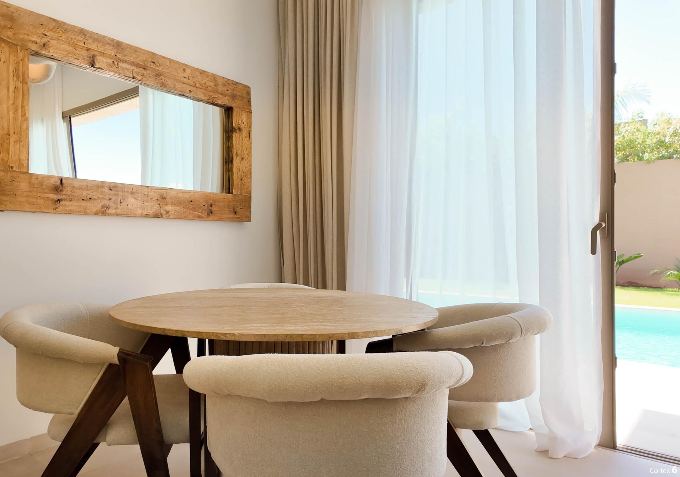 A room with a round wooden table, two beige armchairs, a wooden-framed mirror, and a view of a swimming pool through sheer curtains.