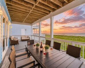 Een overdekt terras met uitzicht op de oceaan bij zonsondergang, met een tafel, stoelen en een fles wijn.