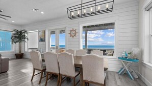 A dining area with a large table and chairs, a hanging light fixture, and a view of the ocean through large windows.