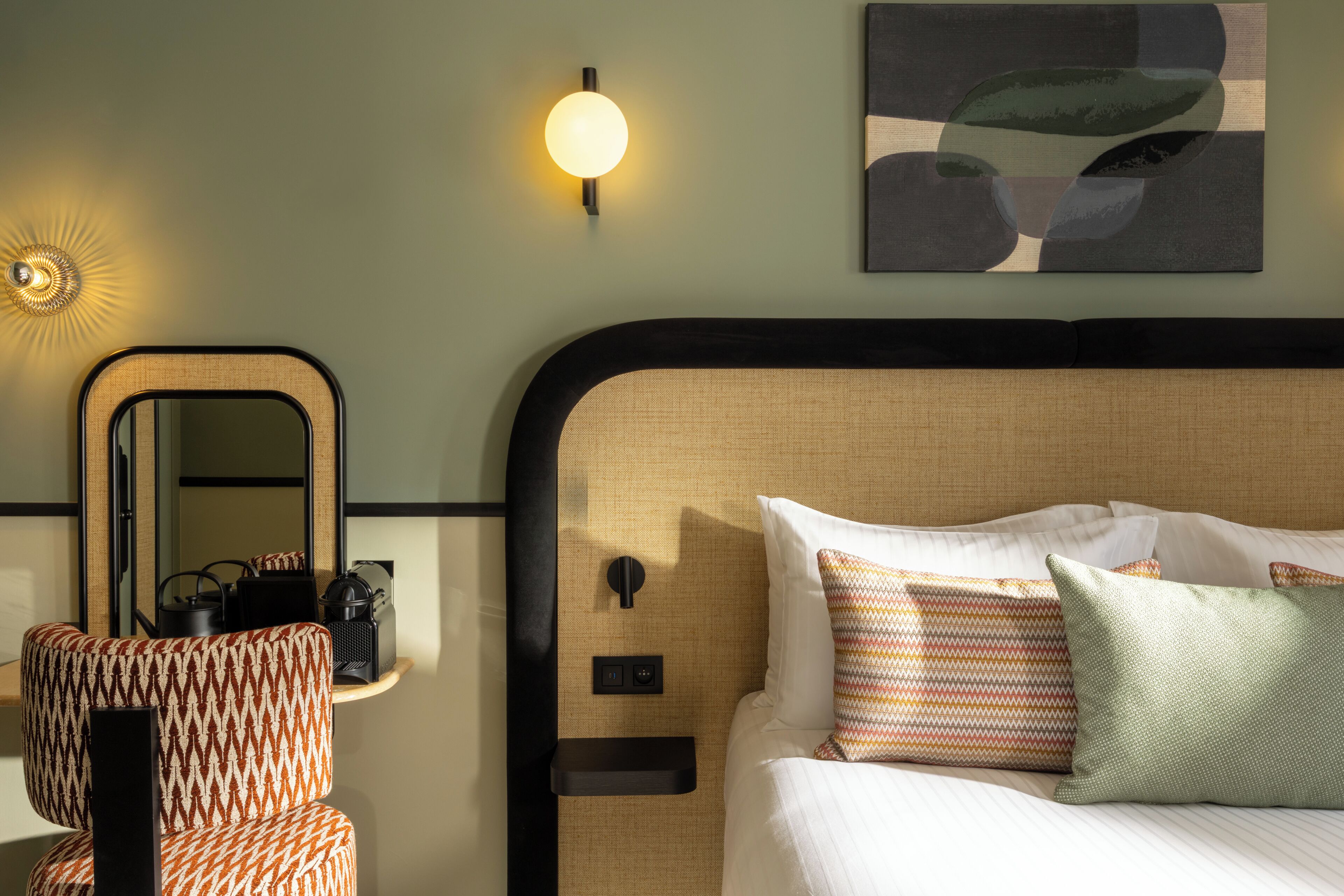A hotel room with a bed, a chair, a nightstand, a wall-mounted lamp, and a painting.