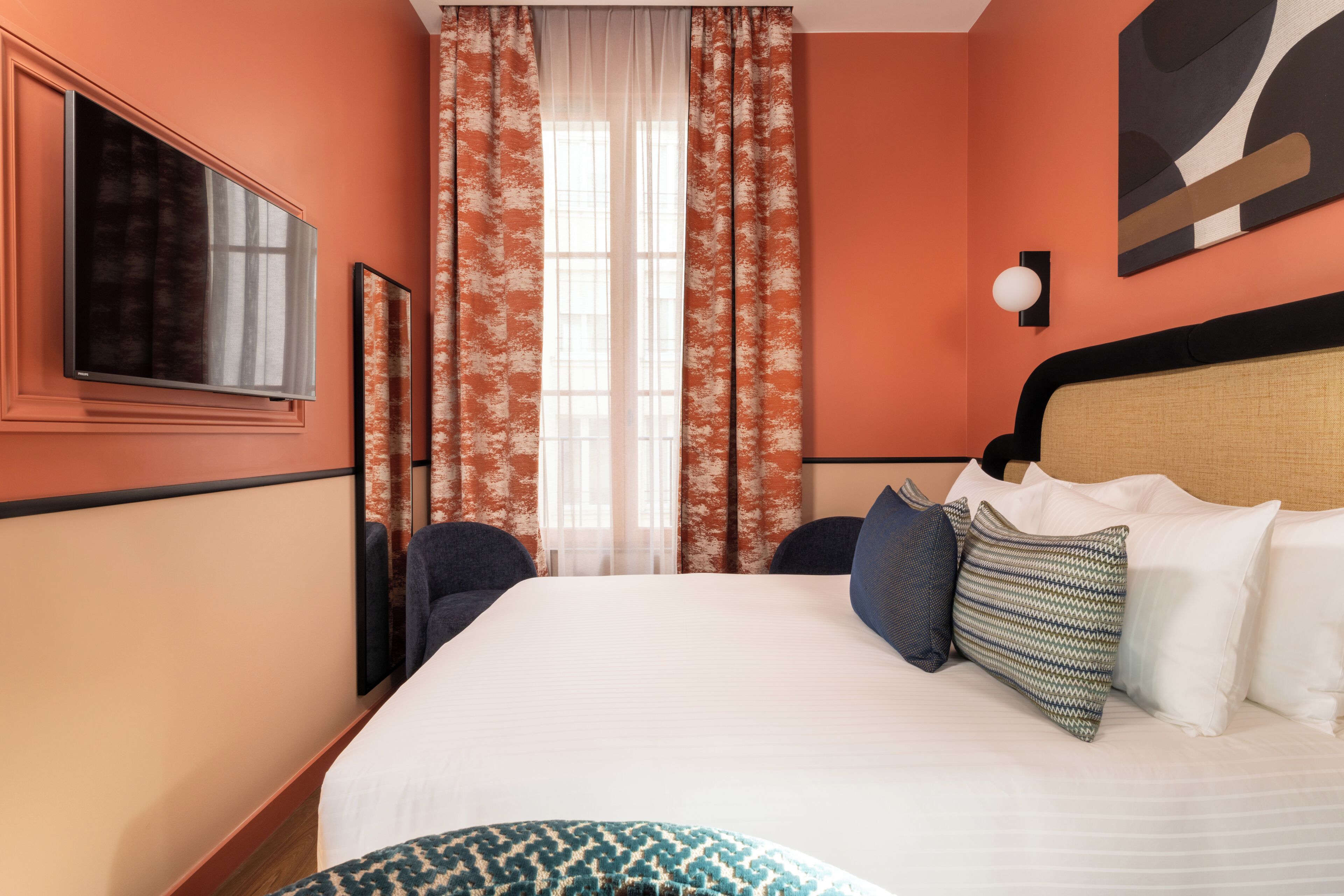 Quarto de hotel com uma cama grande, televisão, janela com cortinas e luminária fixada na parede.