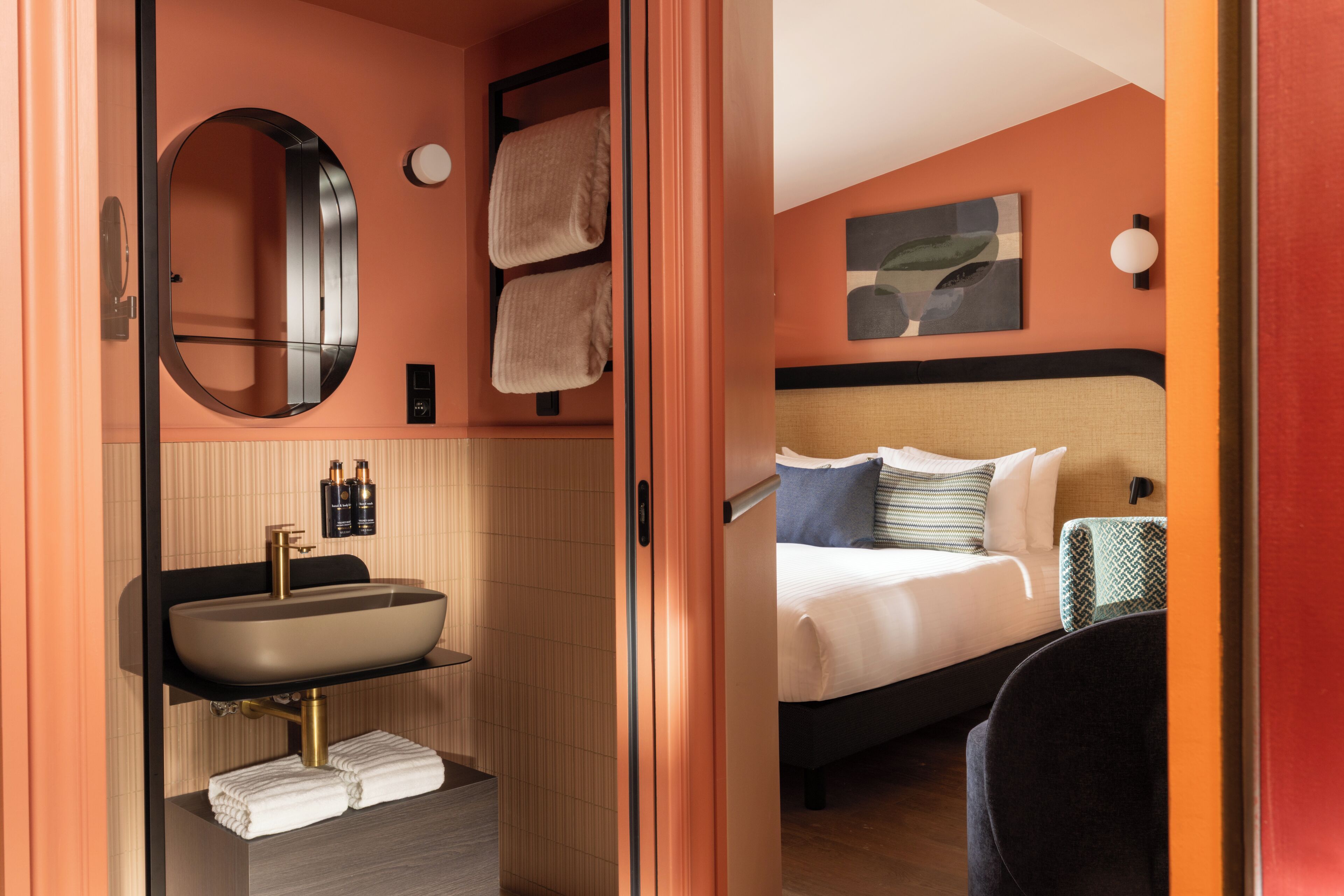 Habitación de hotel con un espejo redondo, un lavamanos, toallas, una cama con almohadas y una obra de arte colgada en la pared.