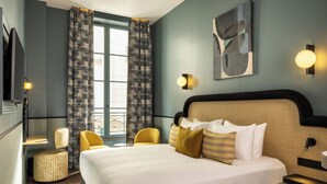 A hotel room with a large bed, a chair, a small table, a wall-mounted TV, and a window with curtains.