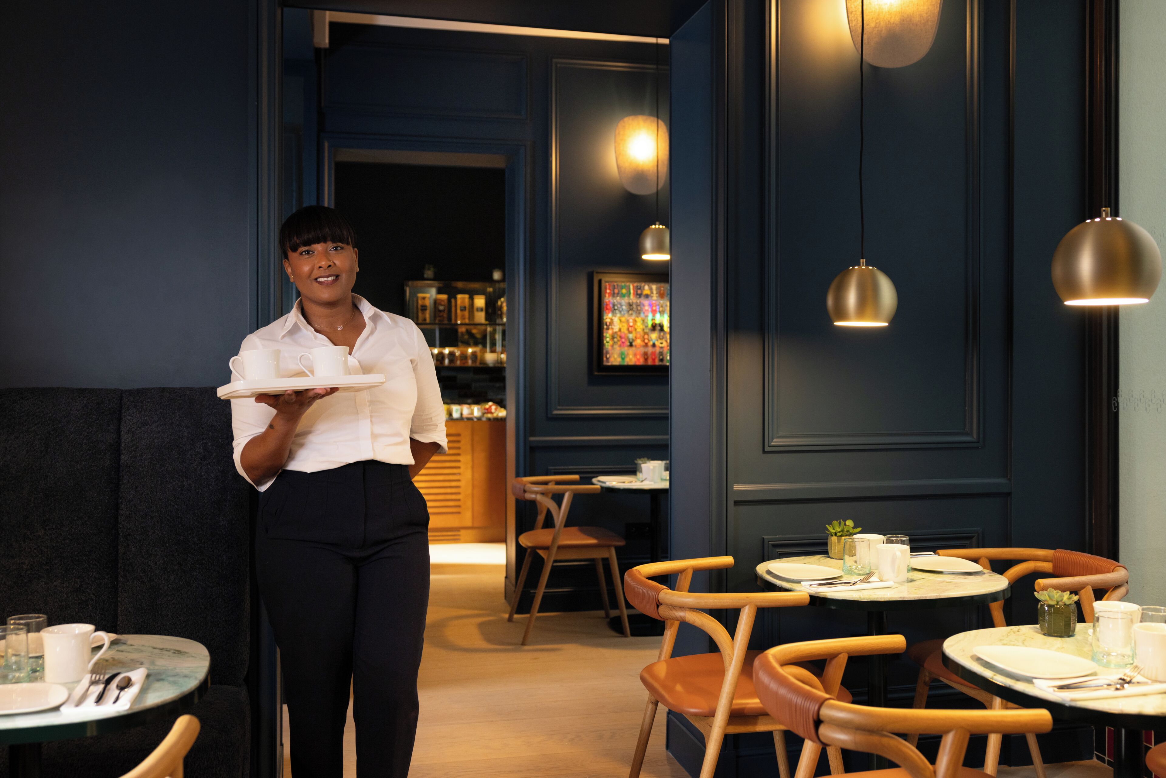 A restaurant interior with wooden chairs, round tables, and a waiter holding a tray.