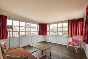 A room with a sofa, a small table, a wicker chair, and large windows with red curtains. - Trastevere Luxury Panoramic Terrace (Roma)