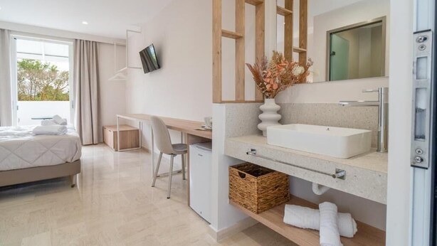 A modern hotel room with a large bed, a desk, a flat-screen TV, a large mirror, a white sink, and a small refrigerator. - Zante Apts (Laganas)