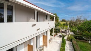 A modern two-story building with a white exterior and red-tiled roof. It features a covered outdoor seating area with tables and chairs, surrounded by well-maintained greenery and a paved pathway. - Zante Apts (Laganas)