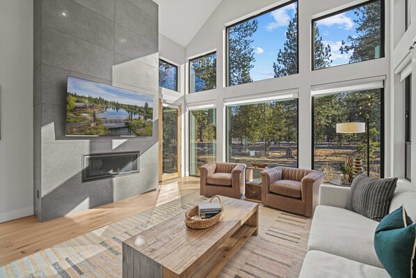 A modern living room with a large TV, comfortable seating, and a view of a lake through large windows. - Modern Caldera Retreat with Access to Exclusive Golf (Sunriver)