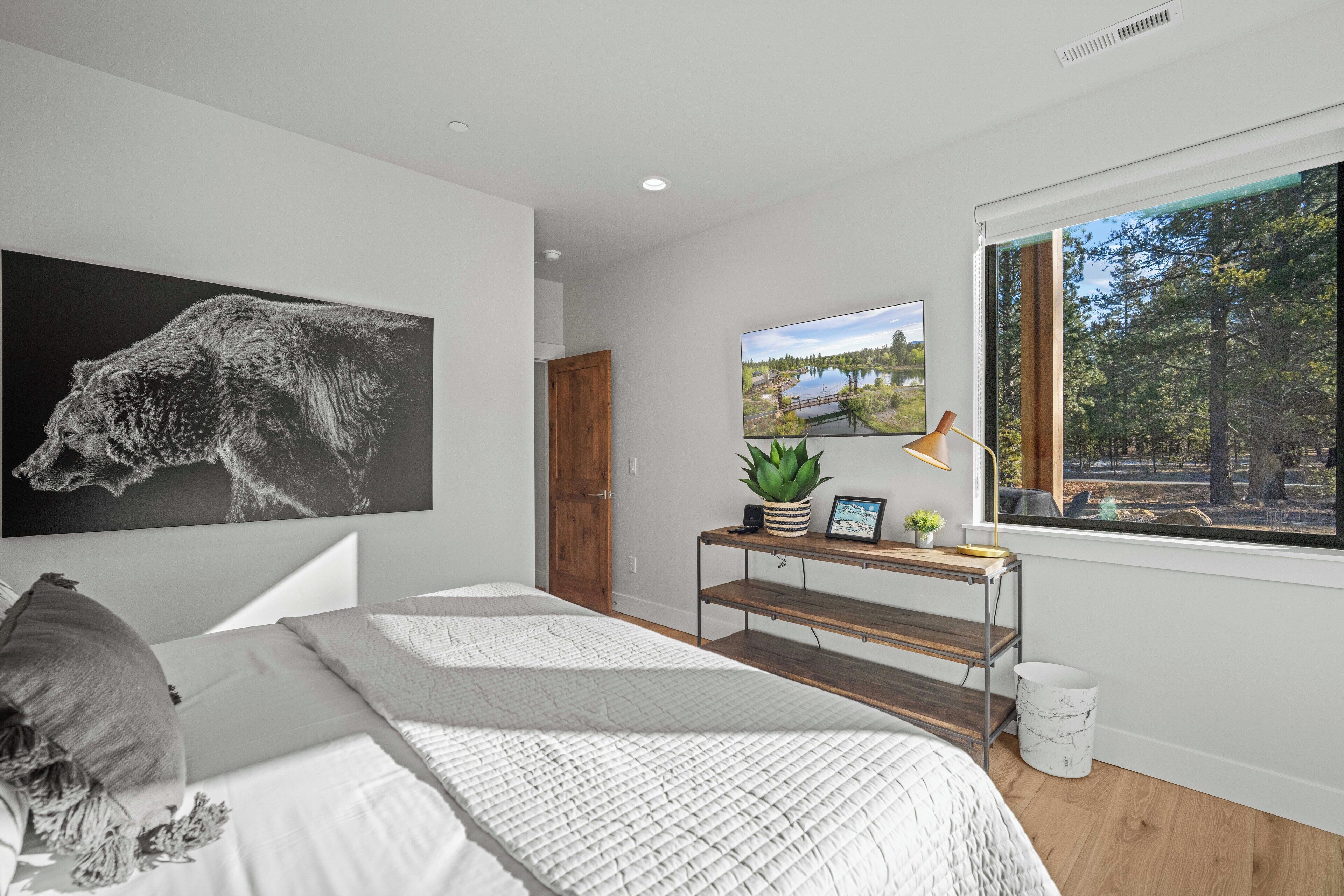 A bedroom with a large bed, a wooden nightstand, a desk with a plant, a window with a view of trees, and a painting on the wall.