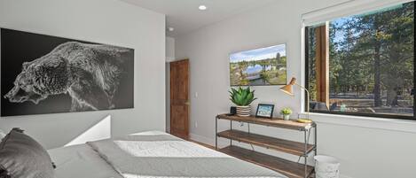 A bedroom with a large bed, a wooden nightstand, a desk with a plant, a window with a view of trees, and a painting on the wall.