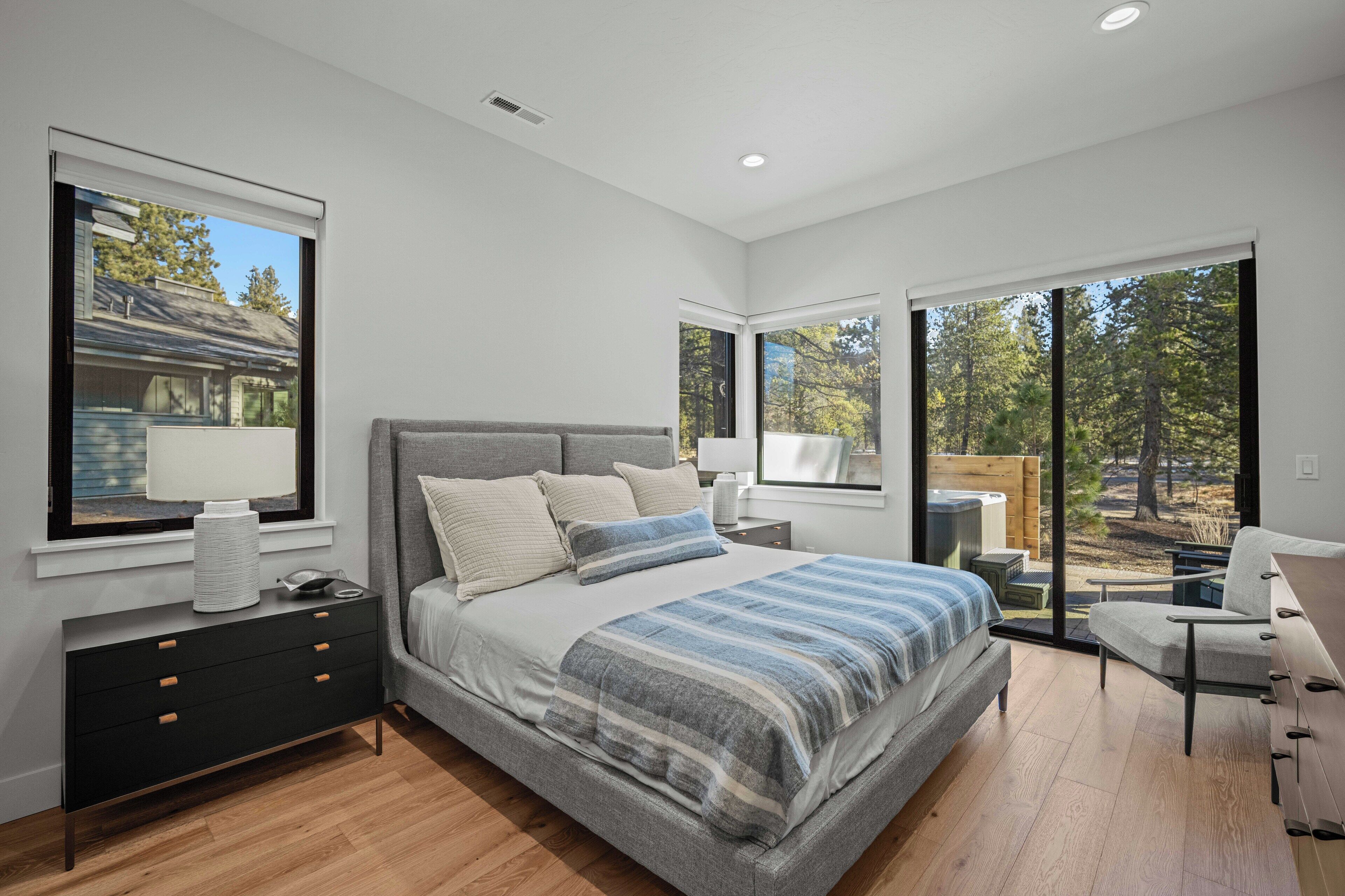 A modern bedroom with a large bed, a nightstand, a dresser, and a chair. There are large windows providing views of the outdoors.