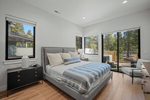 A modern bedroom with a large bed, a nightstand, a dresser, and a chair. There are large windows providing views of the outdoors. - Modern Caldera Retreat with Access to Exclusive Golf (Sunriver)
