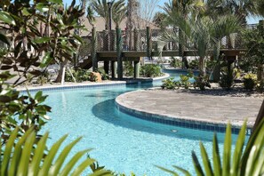 A swimming pool with a curved edge, surrounded by palm trees and a wooden bridge.