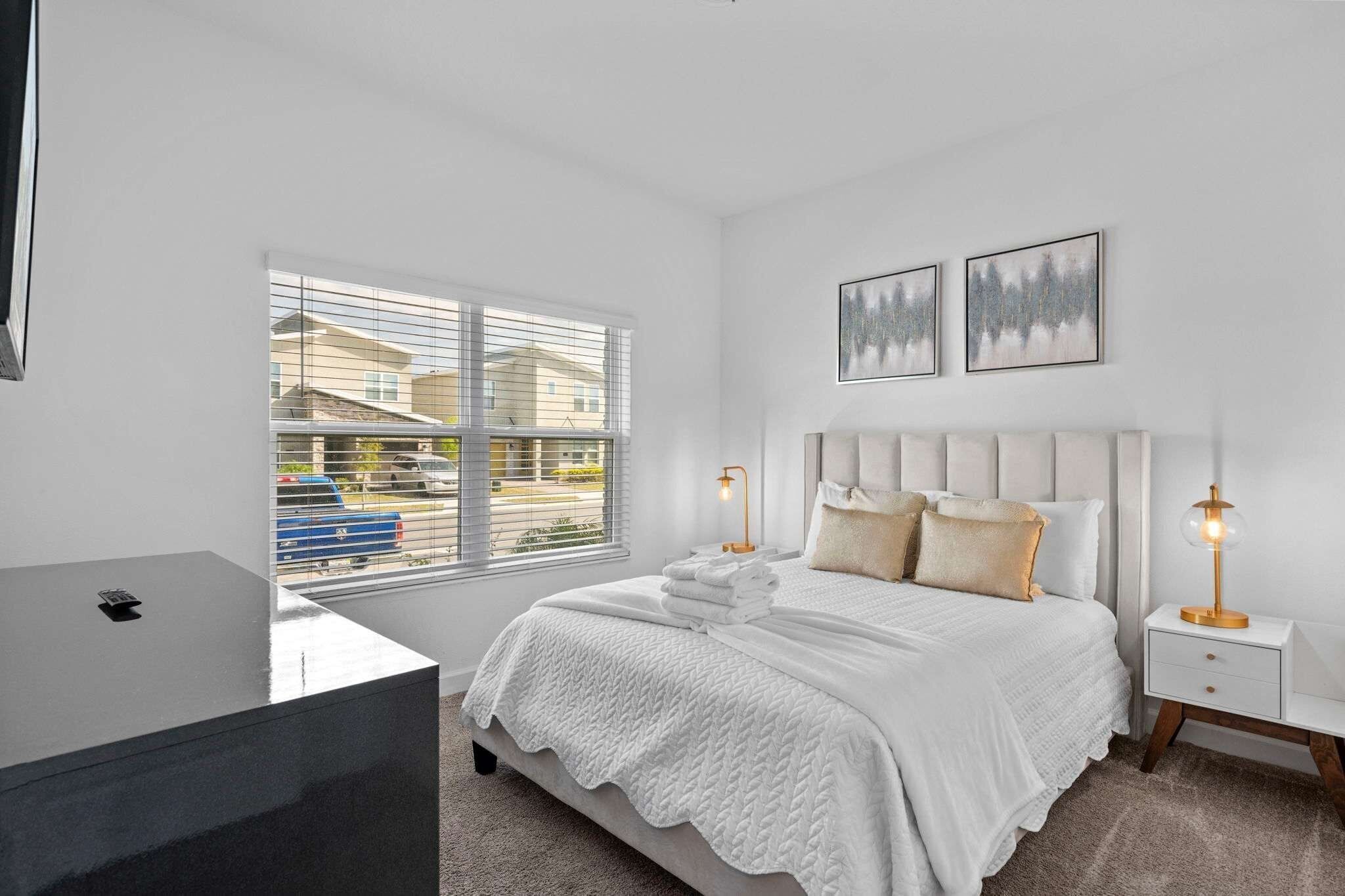 A bedroom with a bed, bedside tables, a window with blinds, and artwork on the wall.