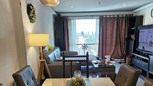Ein modernes Wohnzimmer mit Essbereich, einer Sofagarnitur, einem Fernseher und Blick auf die Stadt durch große Fenster.