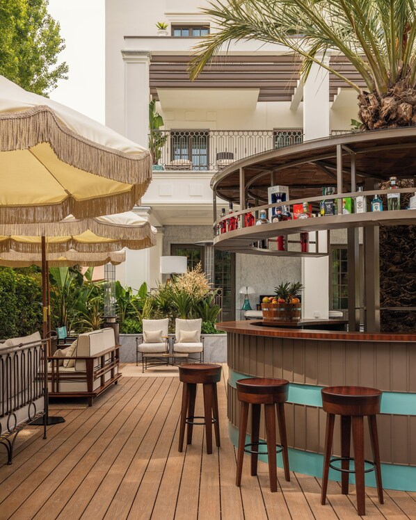 A wooden deck with outdoor seating, a bar area with shelves of bottles, and a view of a building with balconies. - Villa Costanza Boutique Hotel (Forte dei Marmi)