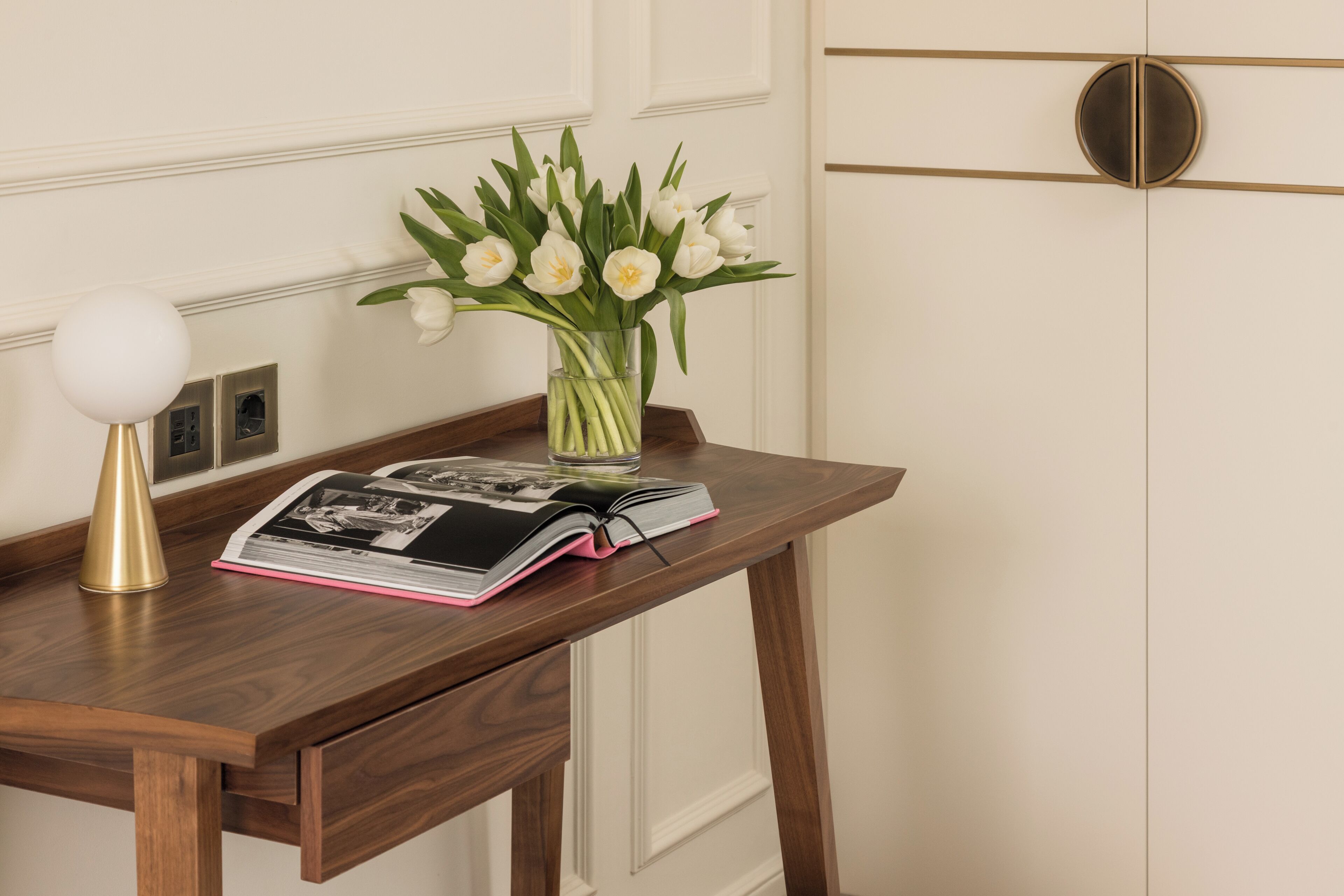 Una mesa de madera con un jarrón de flores blancas, un libro y una lámpara.