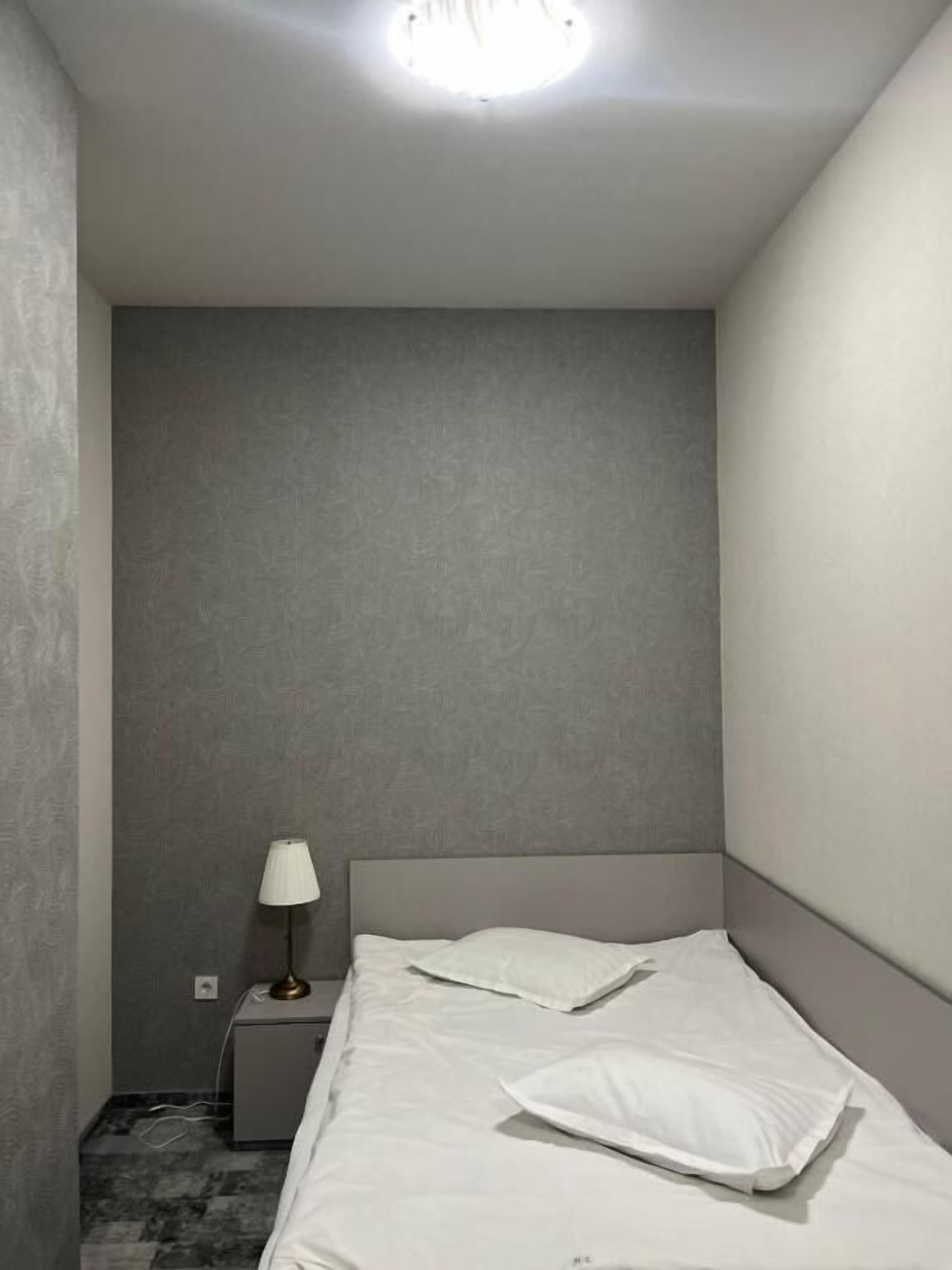 A small, modern bedroom with a single bed, a bedside table with a lamp, and a plain wall.
