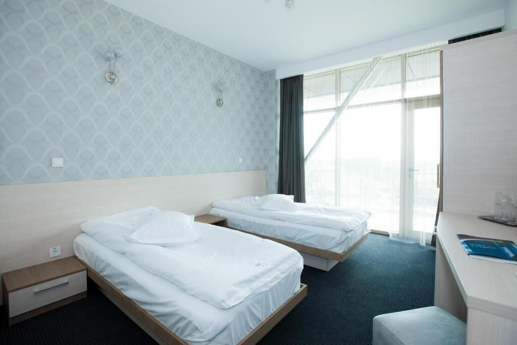 A hotel room with two single beds, a desk, a chair, a small table, and a window with curtains.