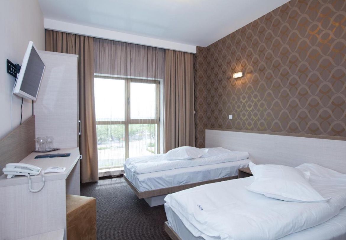 A hotel room with two beds, a desk with a phone, a TV, and a large window with curtains.