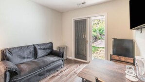 Ein Wohnzimmer mit Ledersofa, Couchtisch aus Holz, Fernseher und einer Schiebetür, die zu einem Garten führt.