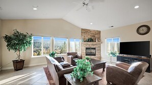 A spacious living room with a stone fireplace, comfortable seating, a large flat-screen TV, and a view of the outdoors through large windows. - Big Yosemite Mansion Large Groups Pinemt Lake (Groveland)