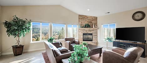 A spacious living room with a stone fireplace, comfortable seating, a large flat-screen TV, and a view of the outdoors through large windows.