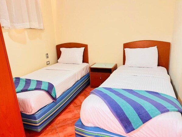 Dos camas individuales con cabeceros de madera, una mesita de noche pequeña y una ventana con cortinas blancas.