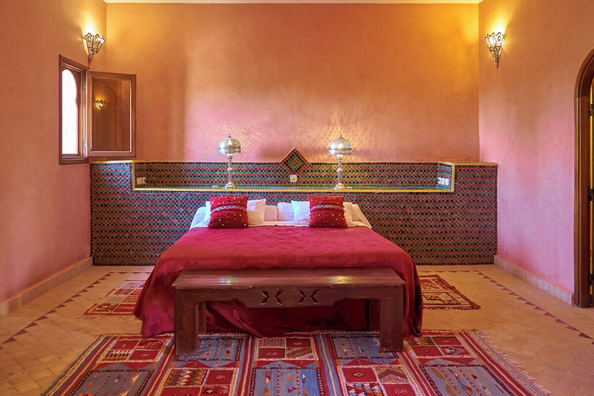 Une chambre avec un grand lit, un banc en bois, des tapis à motifs et un panneau décoratif mural.
