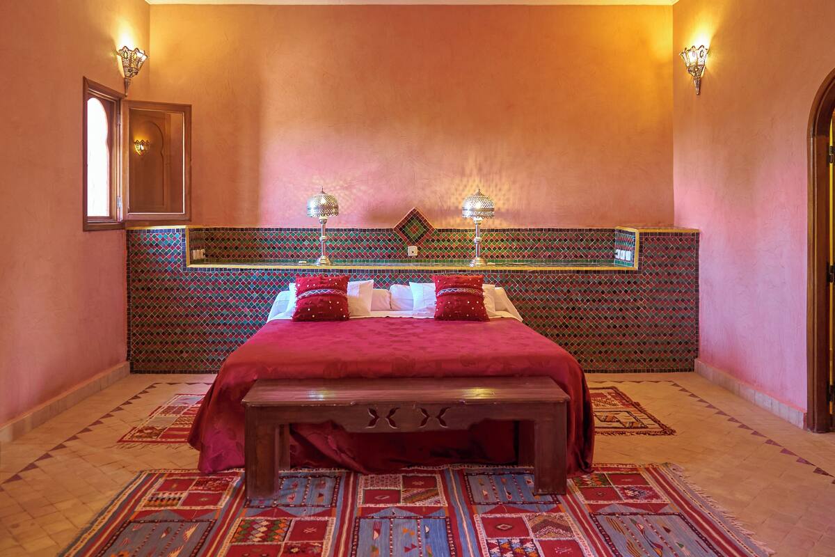 Une chambre avec un grand lit, un banc en bois, des tapis à motifs et un panneau décoratif mural.