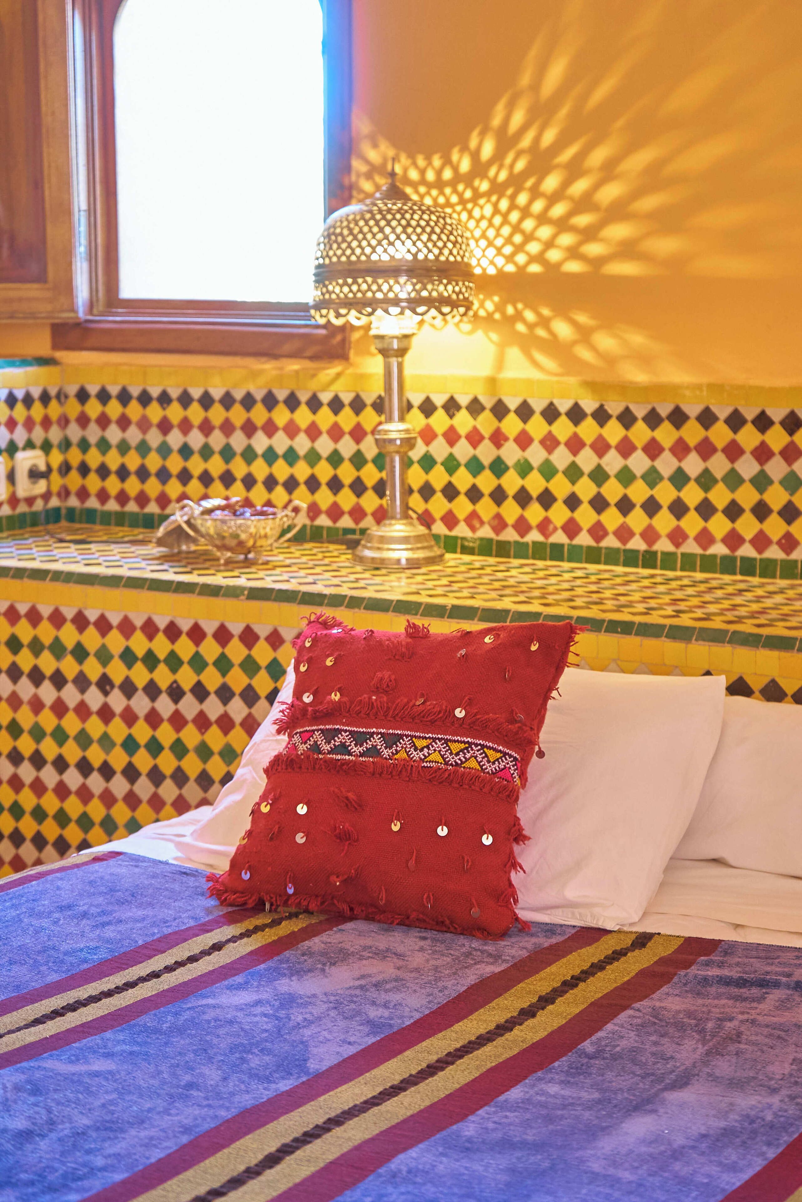 Una habitación con cama, un papel tapiz decorativo, una lámpara y una ventana.