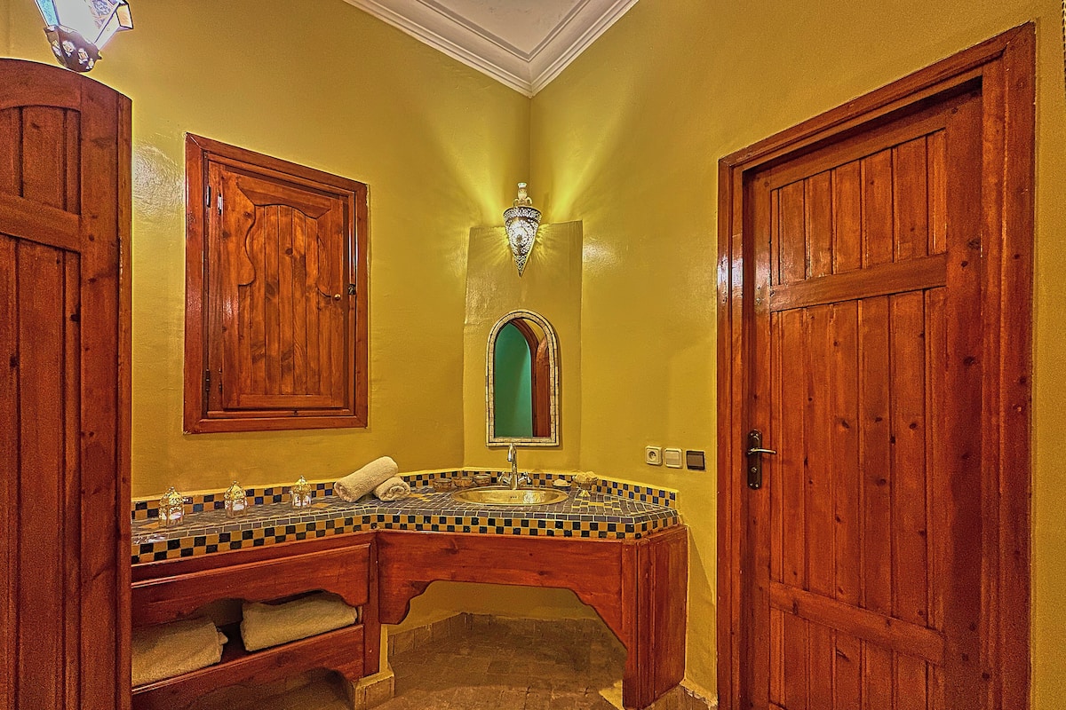 Une salle de bain avec un meuble-lavabo en bois, un évier et un miroir. On y trouve deux portes en bois et un sol carrelé.