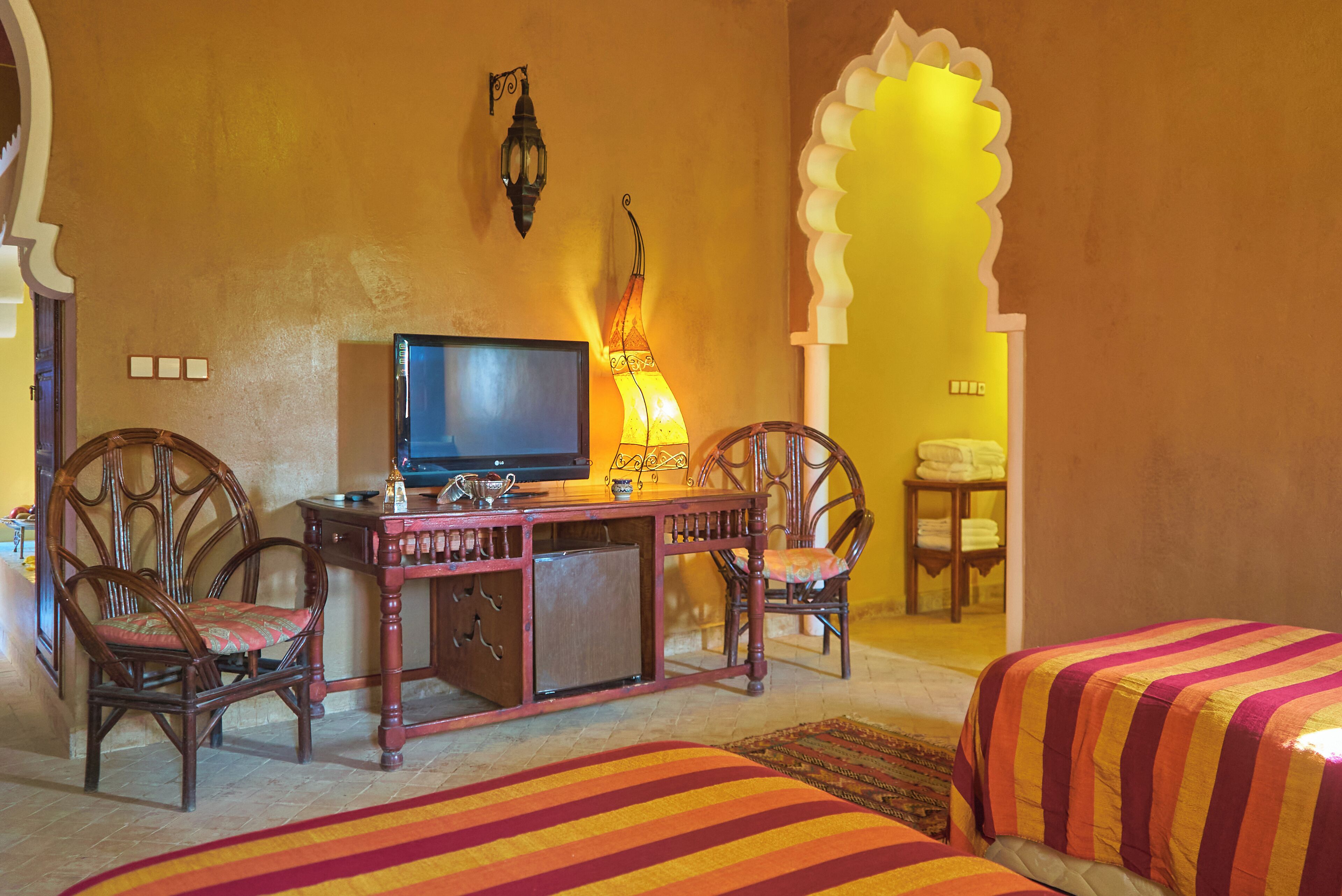 A room with a television, two wooden chairs, a desk, and two beds with striped bedding.