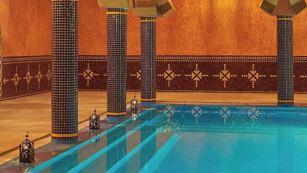 Ein Schwimmbecken mit gefliesten Wänden und Säulen, sowie eine zentrale Fontäne.