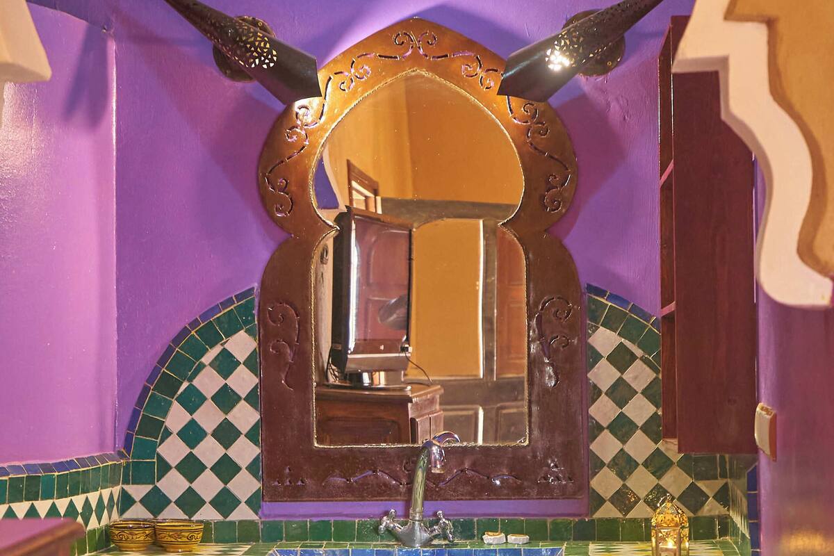 Une salle de bain avec un mur violet, un miroir agrémenté d’éléments décoratifs, un lavabo et un petit tabouret en bois sur lequel sont posées des serviettes.